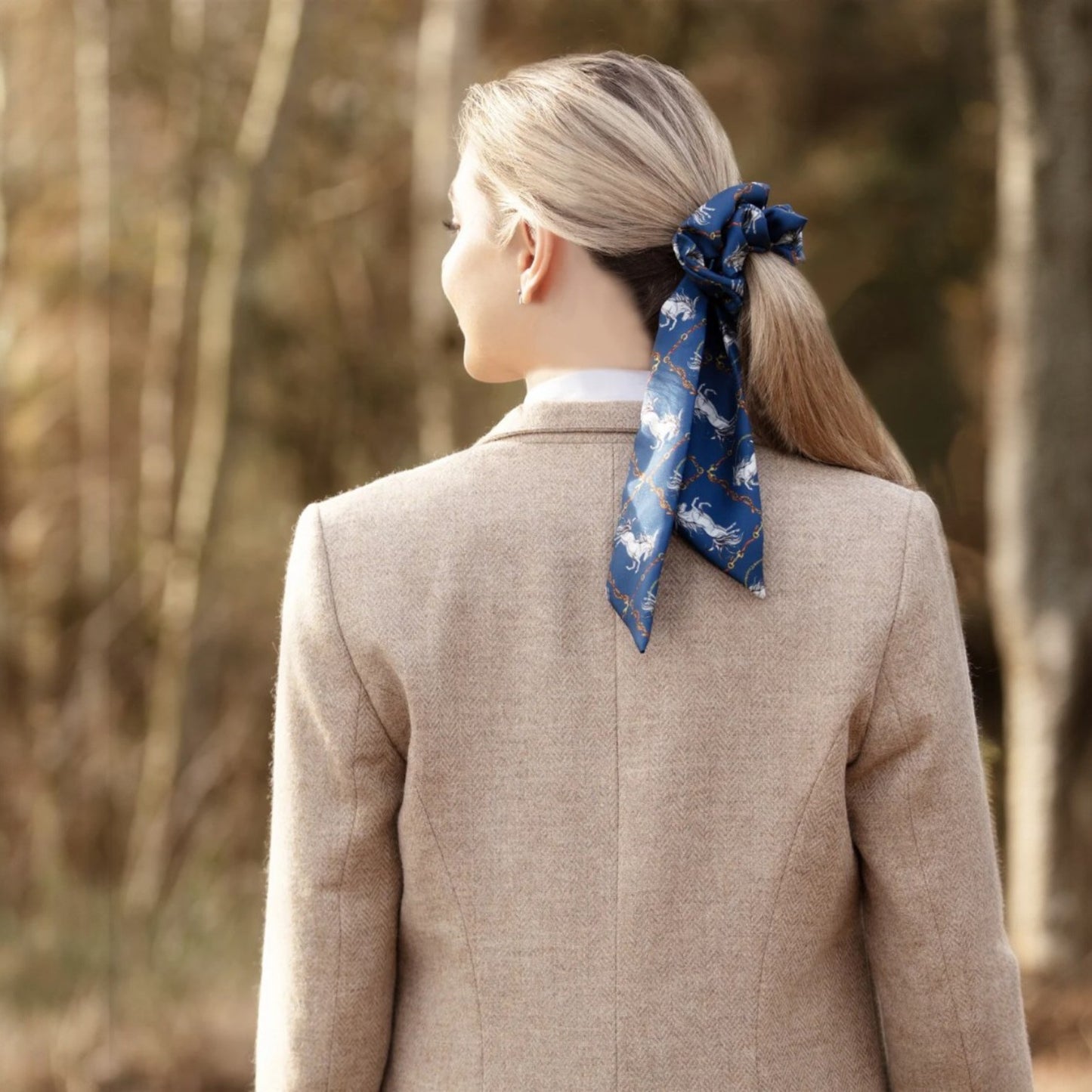 Person wearing a beige coat with a blue scarf tied in their hair, standing in a forest.