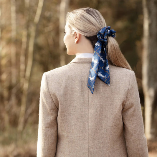 Person wearing a beige coat with a blue scarf tied in their hair, standing in a forest.