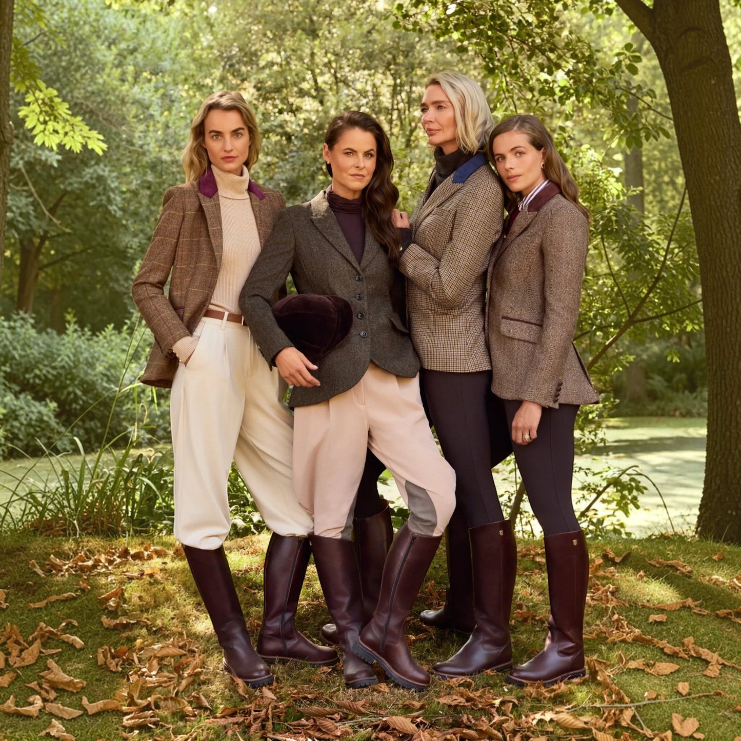 Four women posing together outdoors in a wooded setting, wearing coordinated equestrian-style outfits with tailored jackets, tall boots, and riding pants.