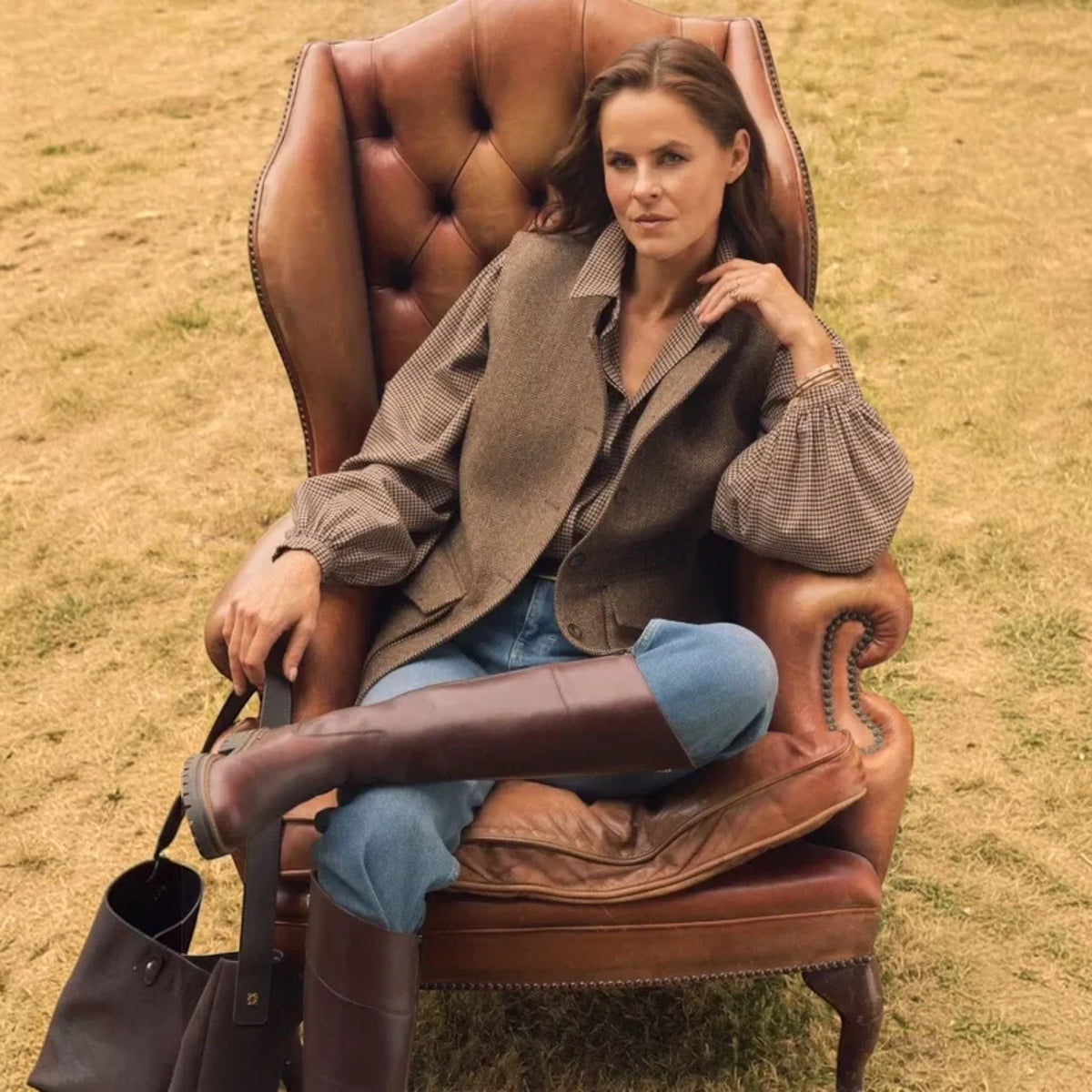 A woman sits in a brown leather wingback armchair outdoors on dry grass, wearing a brown textured jacket, blue jeans, and dark brown knee-high boots. A dark tote bag rests beside her on the chair.
