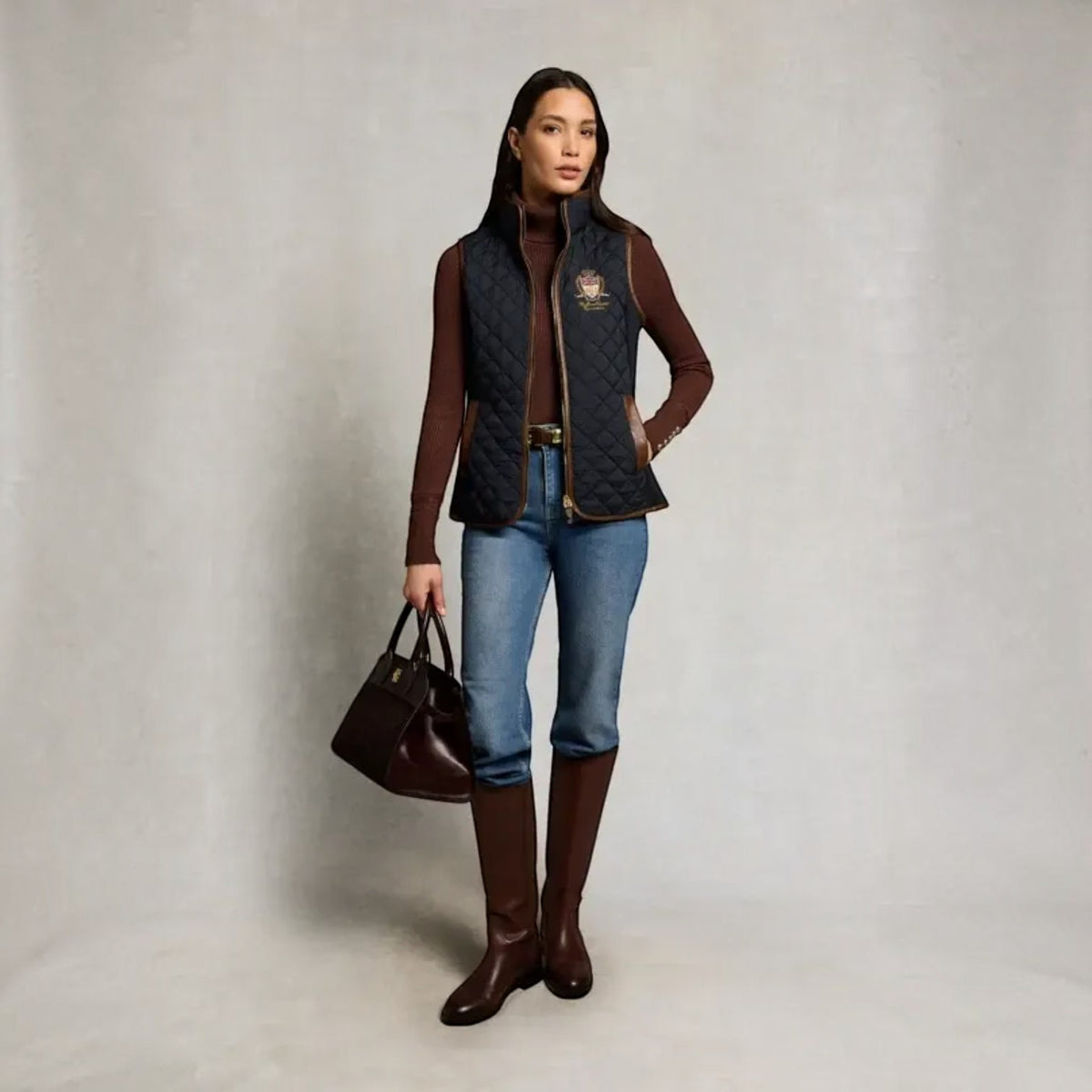 A woman stands against a light gray studio backdrop wearing a dark quilted zip-front vest over a brown turtleneck, blue jeans, and brown knee-high boots, while holding a dark brown handbag.