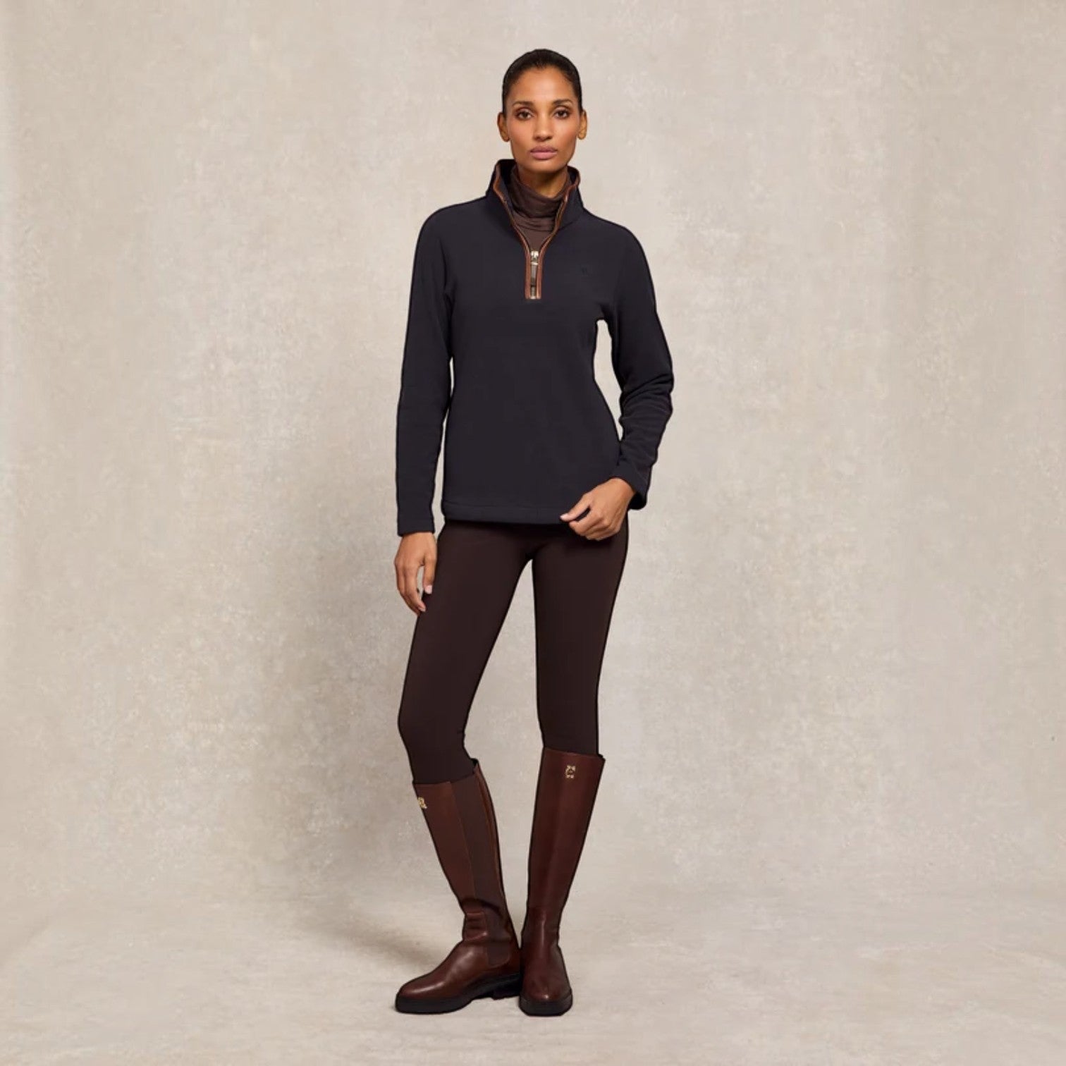 A woman stands against a textured beige studio backdrop wearing a dark navy quarter-zip long-sleeve top over a brown mock-neck layer, dark brown fitted leggings, and dark brown riding boots.