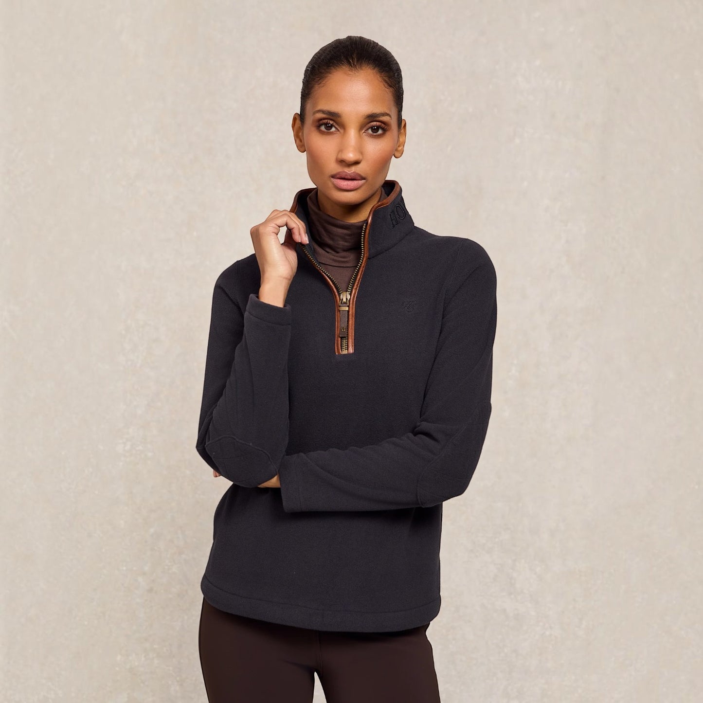 A woman modeling a dark charcoal quarter-zip fleece pullover with a brown collar lining and a brown zipper pull, paired with dark brown leggings, against a textured beige background.