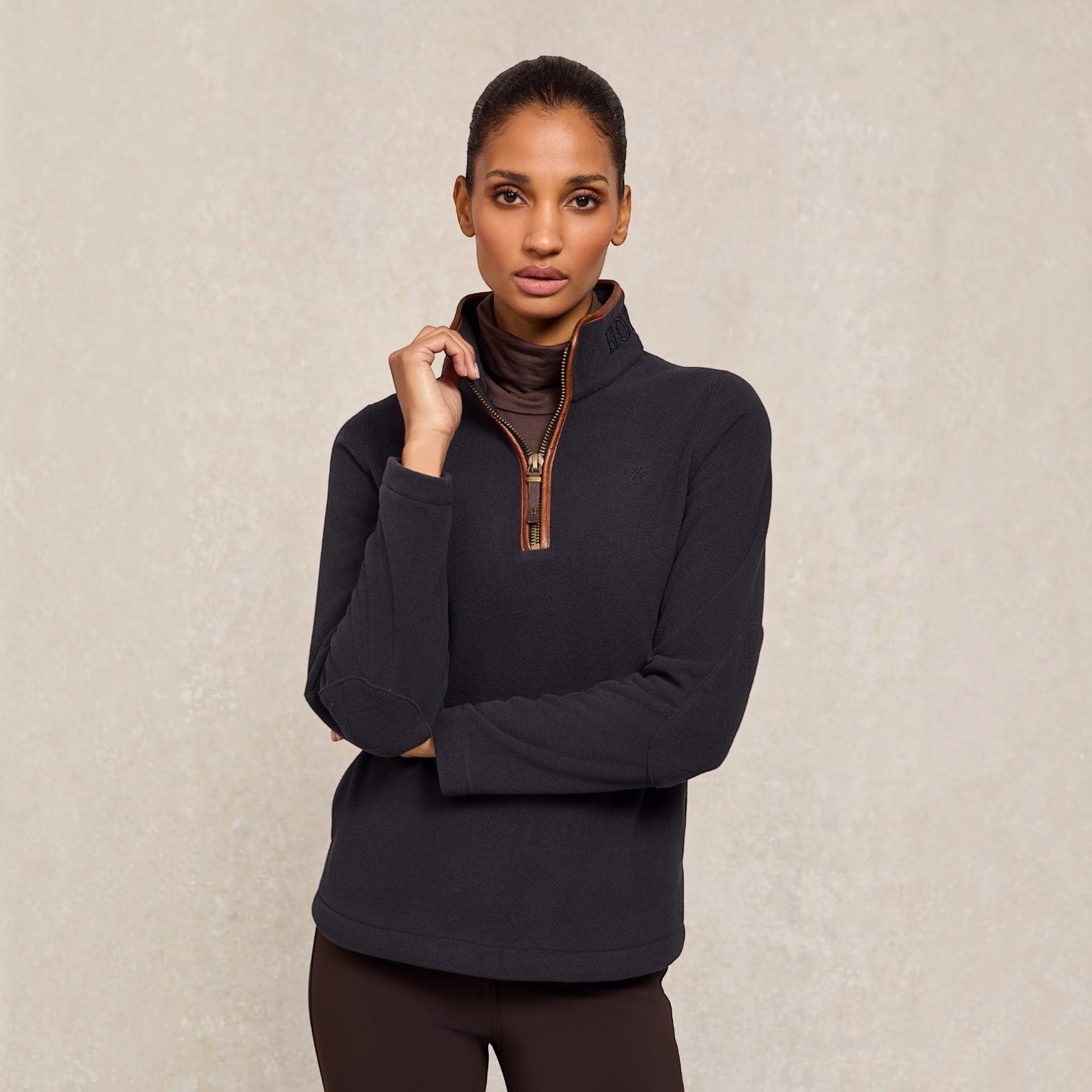 A woman modeling a dark charcoal quarter-zip fleece pullover with a brown collar lining and a brown zipper pull, paired with dark brown leggings, against a textured beige background.