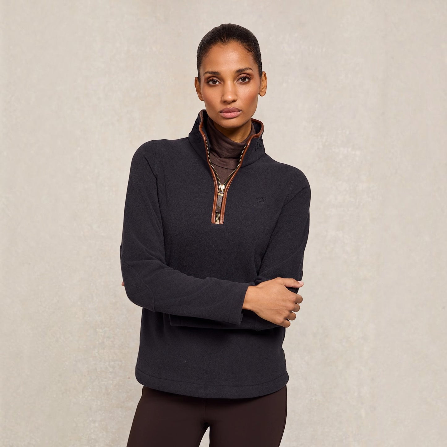 A woman modeling a dark charcoal quarter-zip pullover with brown trim over a brown turtleneck, set against a textured light beige background.
