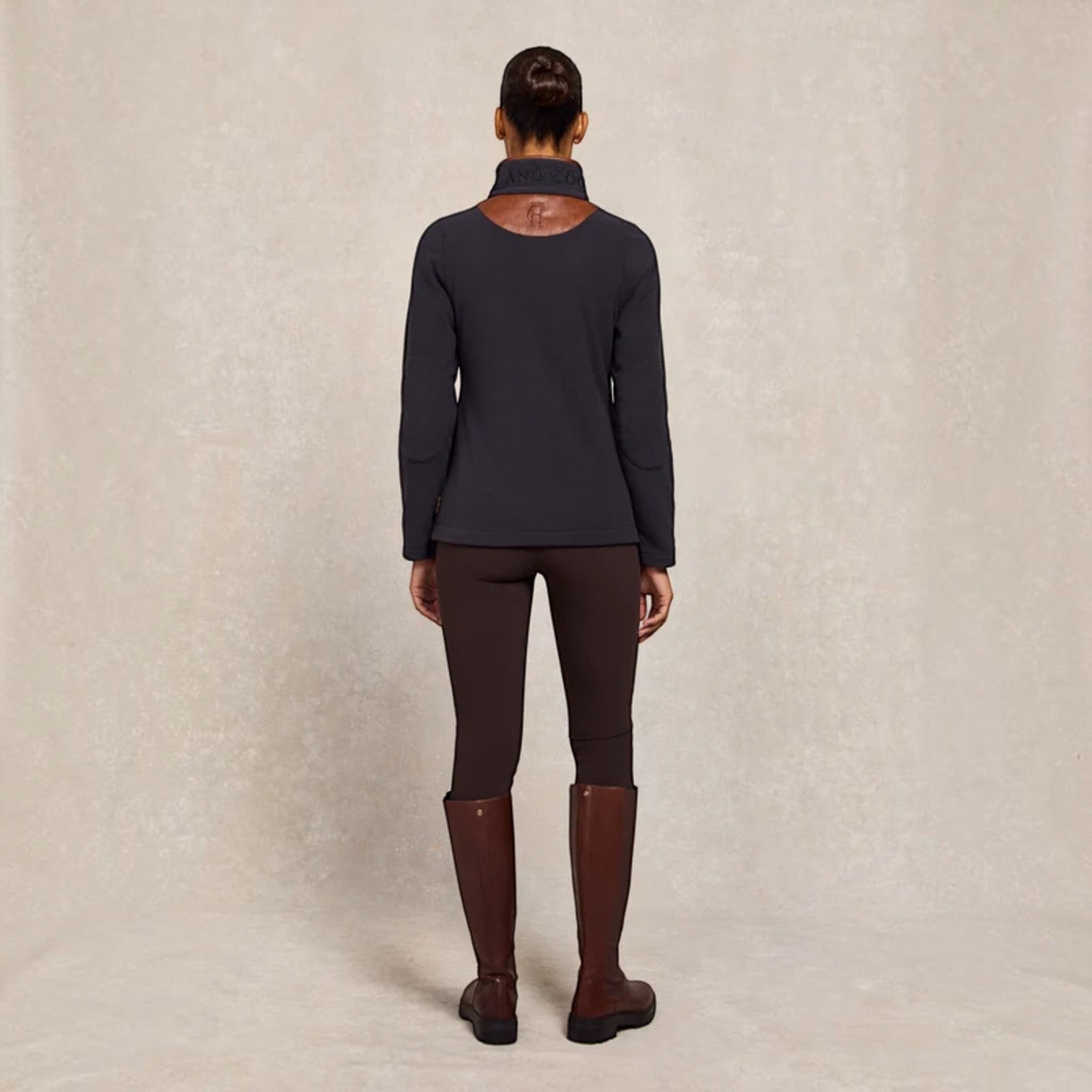 A model shown from the back wearing a dark charcoal long-sleeve equestrian shirt, brown riding breeches, and tall brown riding boots against a textured beige backdrop.
