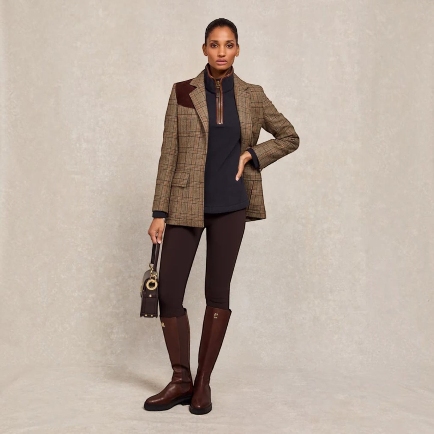 A model stands against a textured beige studio backdrop wearing a brown and beige plaid blazer with dark brown leather shoulder patches, a navy mock-neck top, dark brown leggings, and dark brown riding boots. She holds a small dark brown handbag.
