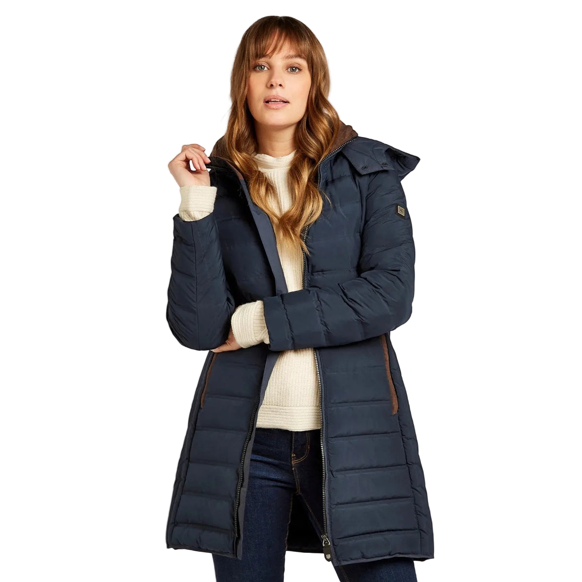A woman wearing a long navy blue quilted puffer coat over a cream ribbed sweater and dark jeans, standing against a light gray studio background.