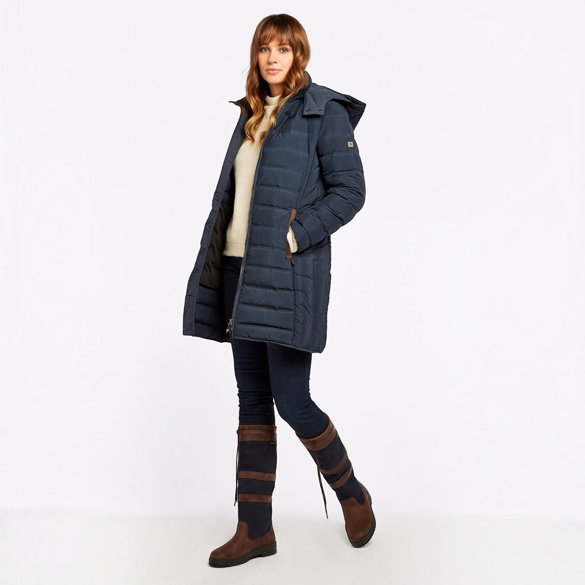 A woman modeling a long navy blue puffer coat with a hood, worn over a cream top and dark pants, paired with brown knee-high boots, against a plain white studio background.