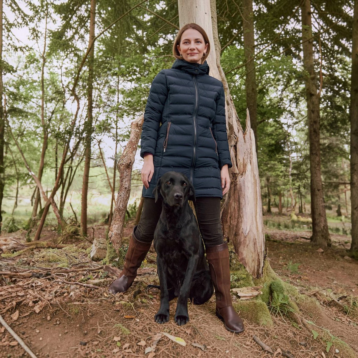 A woman wearing a dark navy puffer coat, black leggings, and brown boots stands in a forest with a black dog sitting in front of her.