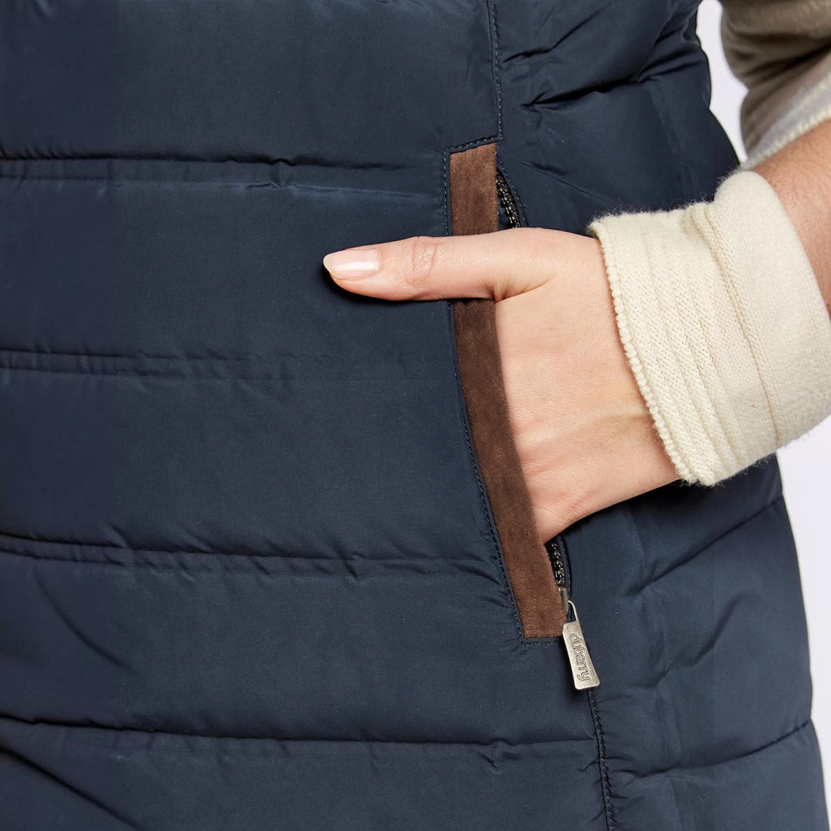 Close-up of a person wearing a dark navy puffer vest with a side pocket, showing a hand in the pocket and a cream ribbed knit sleeve.