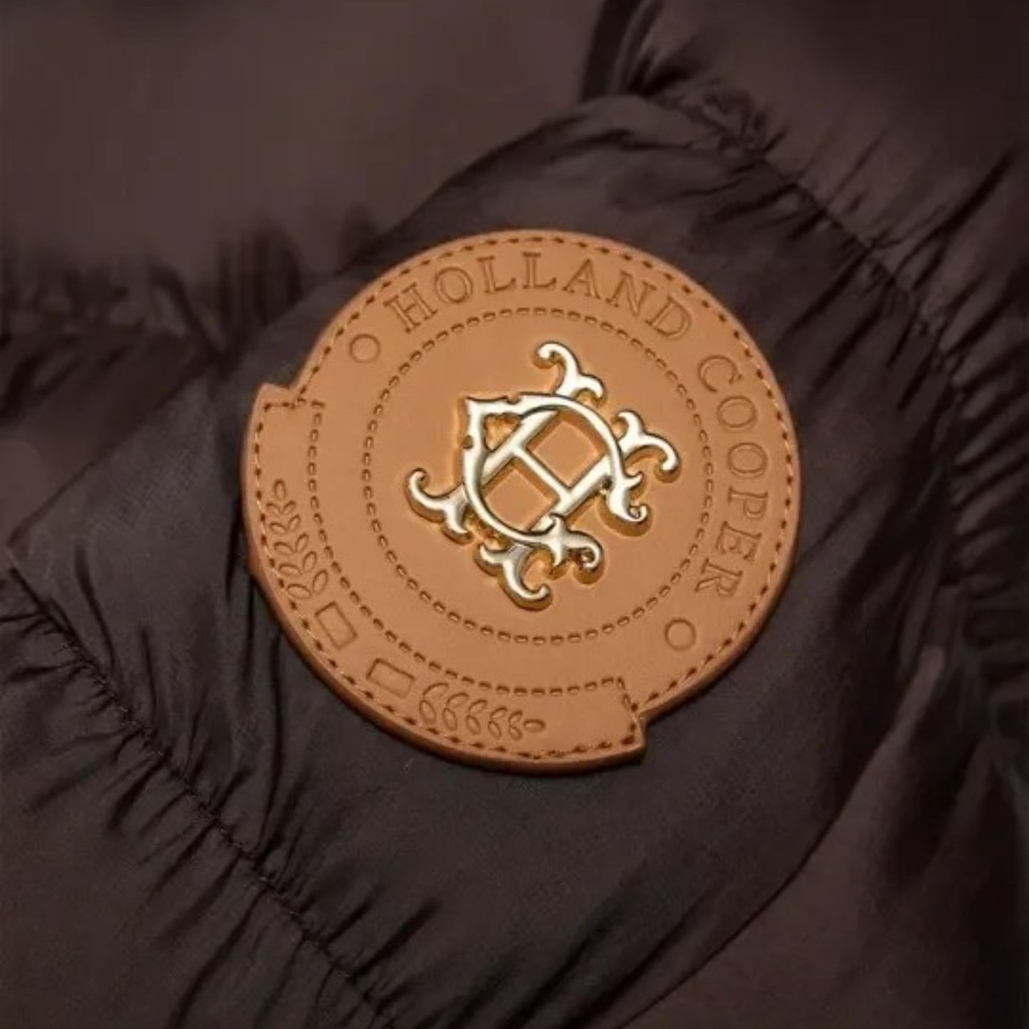 A close-up of a black puffer jacket sleeve featuring a circular tan leather patch with a gold-tone metal emblem and the text 'HOLLAND COOPER'.