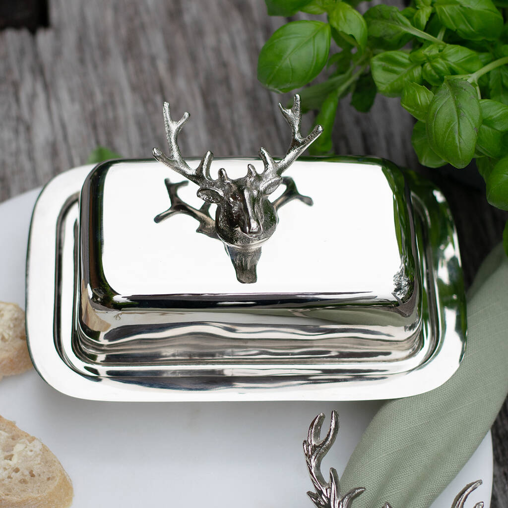 Silver butter dish with deer head design on a table with bread and greenery.