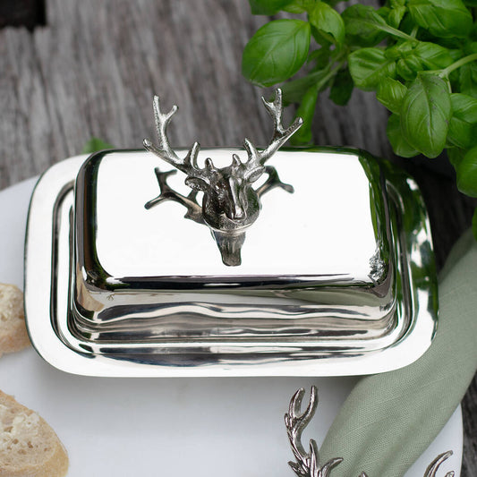 Silver butter dish with deer head design on a table with bread and greenery.