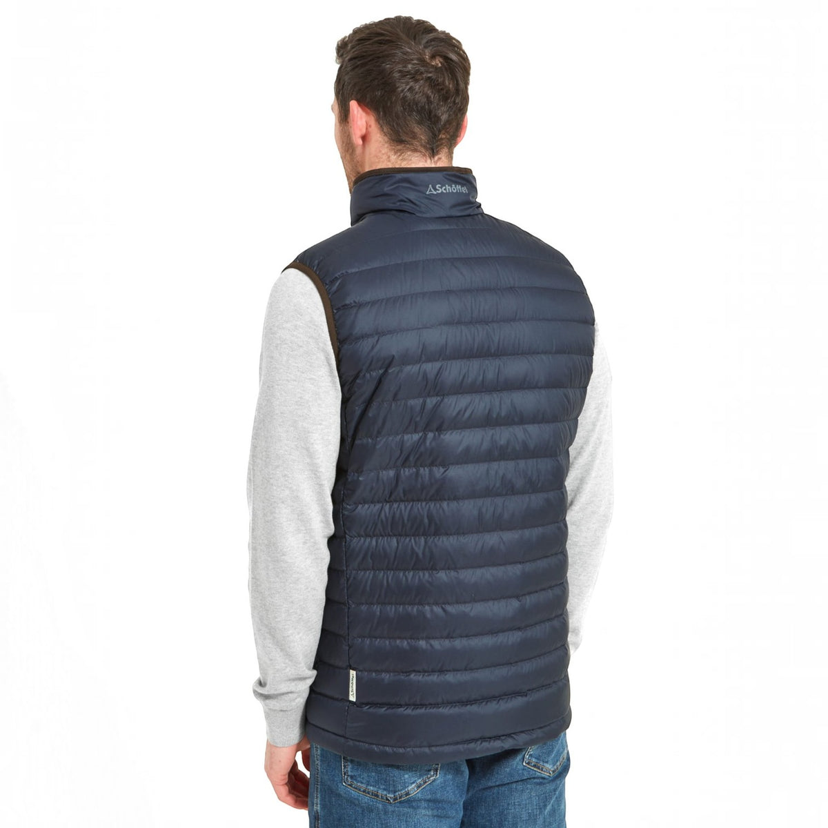 A man viewed from behind wearing a navy blue puffer vest over a light gray long-sleeve shirt and blue jeans, against a plain white background.