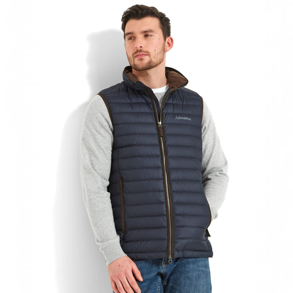 A man wearing a dark navy hooded puffer vest with brown trim over a light gray long-sleeve shirt and blue jeans, standing against a white studio background.