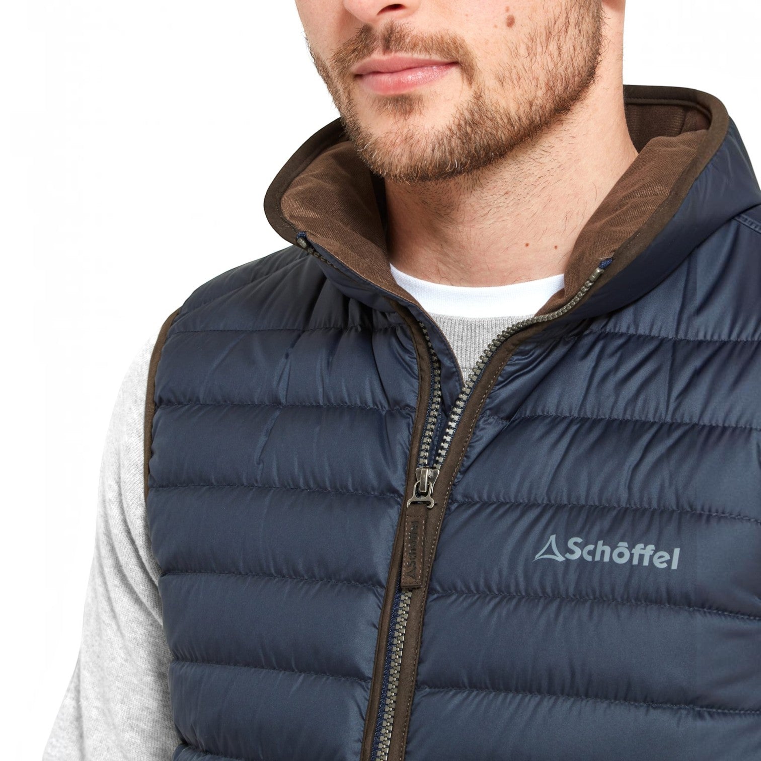 A close-up of a man wearing a navy blue Schöffel puffer vest with a brown interior lining and a white crewneck shirt underneath, against a plain white background.