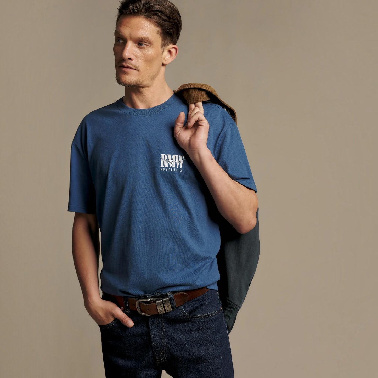 A man wearing a blue ribbed short-sleeve t-shirt with a white 'RMW' logo on the chest, paired with dark jeans and holding a brown leather jacket over his shoulder, set against a solid beige studio background.
