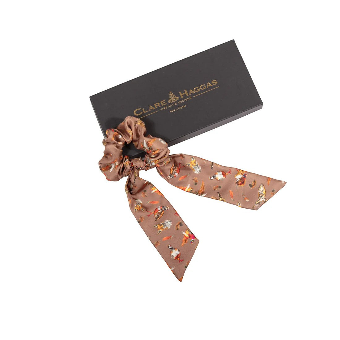 Brown hair scrunchie with decorative ribbon in front of a Clare Haggas box on a white background