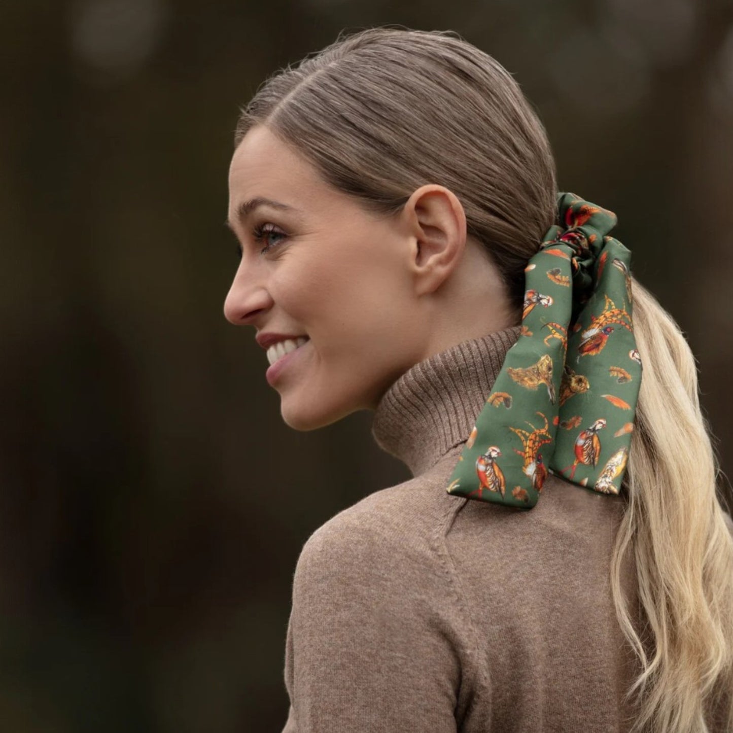 Woman with a patterned scrunchie in her hair, wearing a brown sweater outdoors.